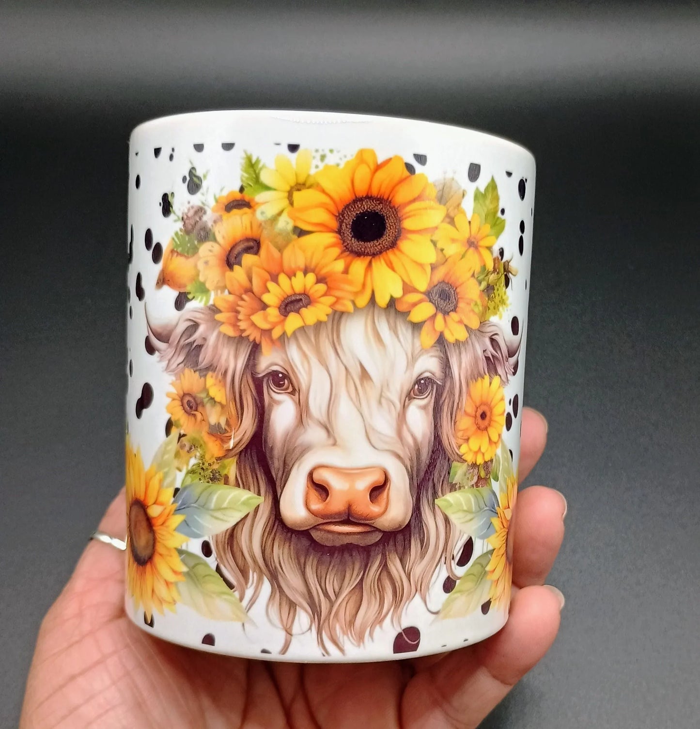White ceramic mug with detailed illustration of a cow wearing a sunflower crown, held in hand