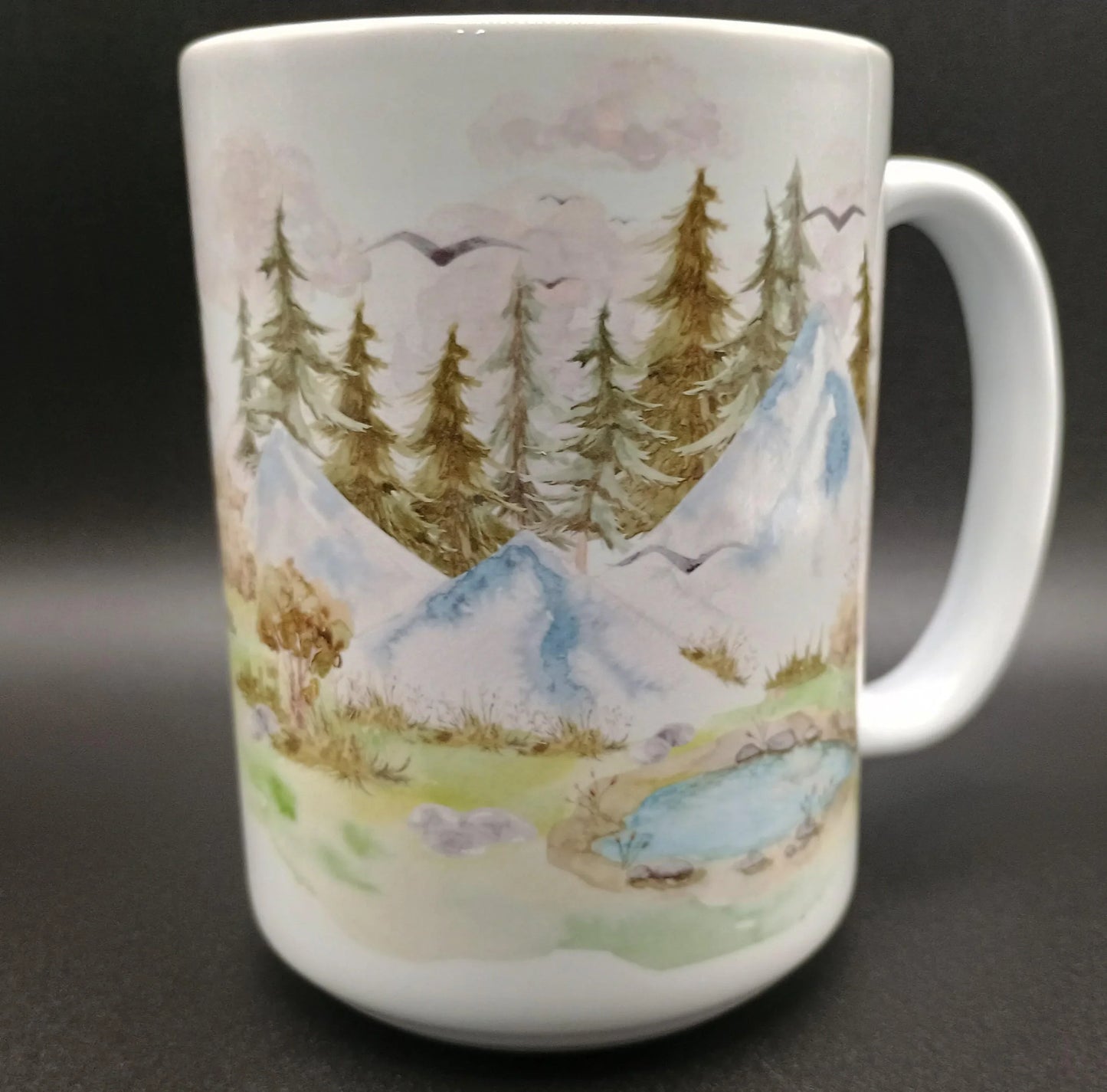 White ceramic mug with hand-painted mountain landscape, pine trees, birds, and a small pond