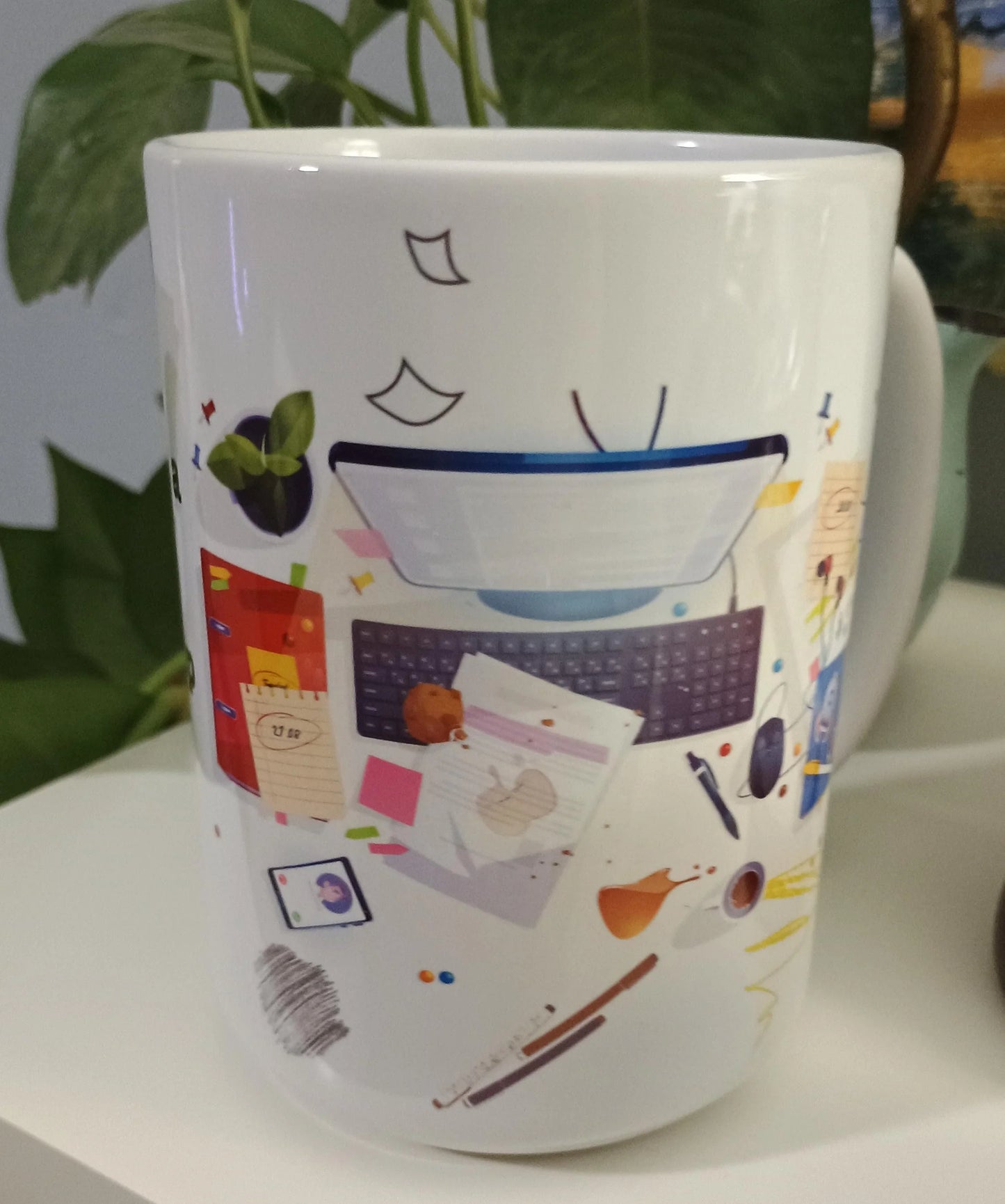 White mug with colorful illustration of a cluttered desk including keyboard, papers, coffee cup, plant, and stationery