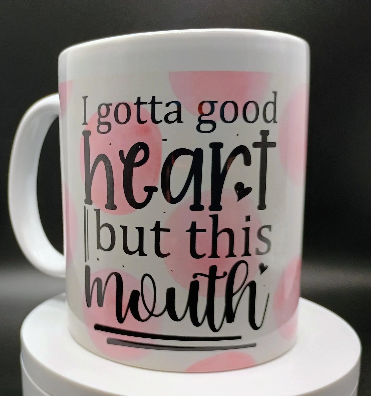 White ceramic mug with pink spots and black text reading 'I gotta good heart but this mouth'