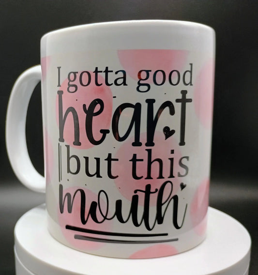 White ceramic mug with pink spots and black text reading 'I gotta good heart but this mouth'