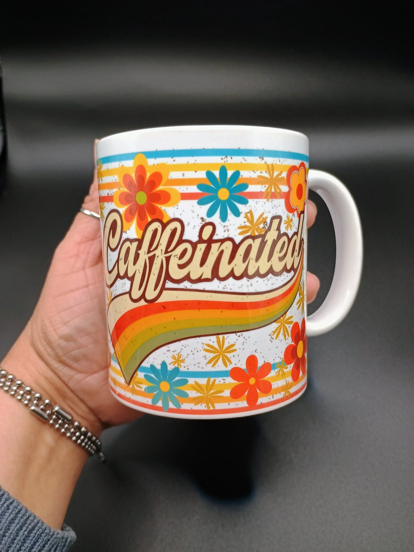 Hand holding white mug with colorful retro floral design and word 'Caffeinated' on black background