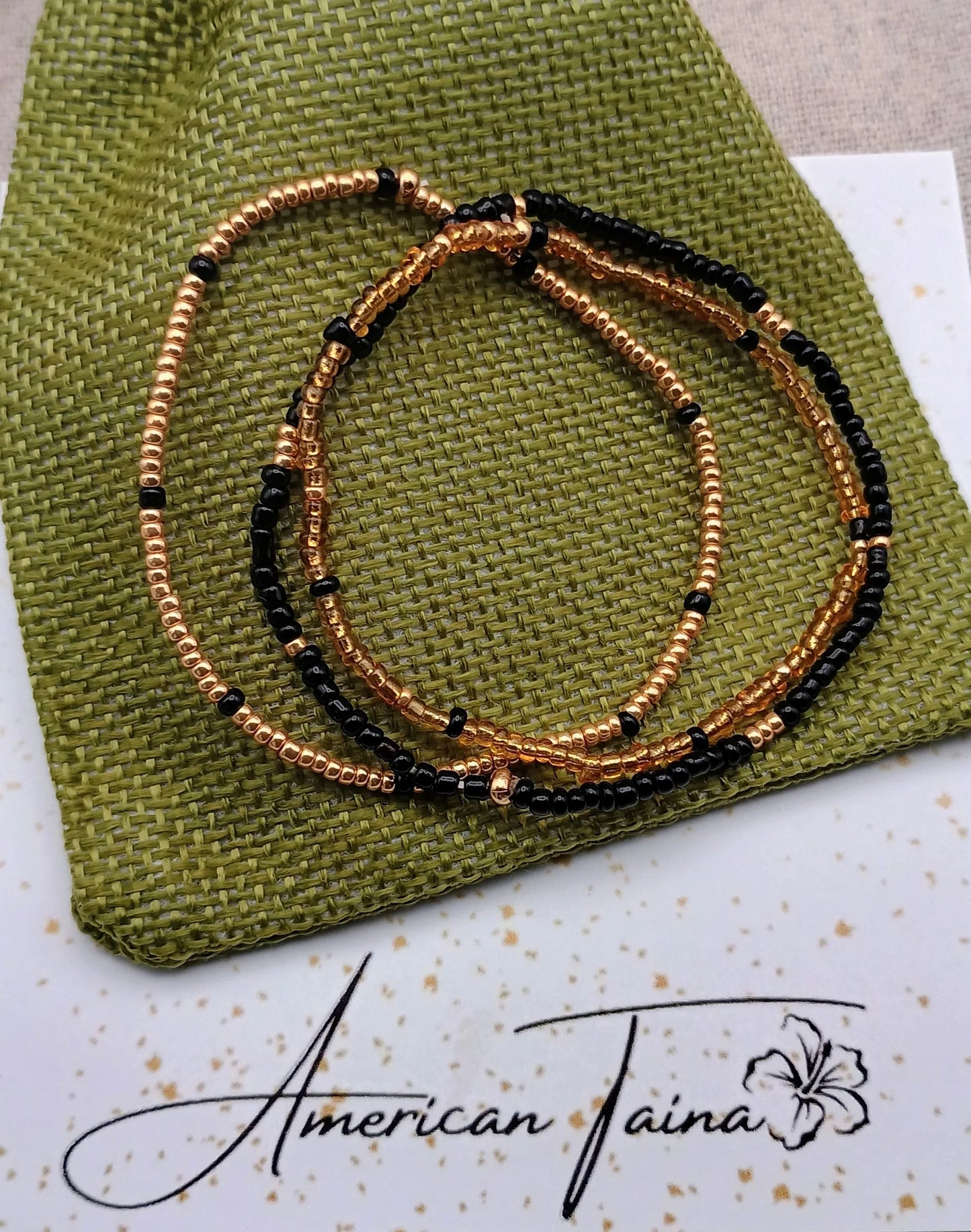 Gold and black beaded bracelets on green textured fabric with American Taina logo