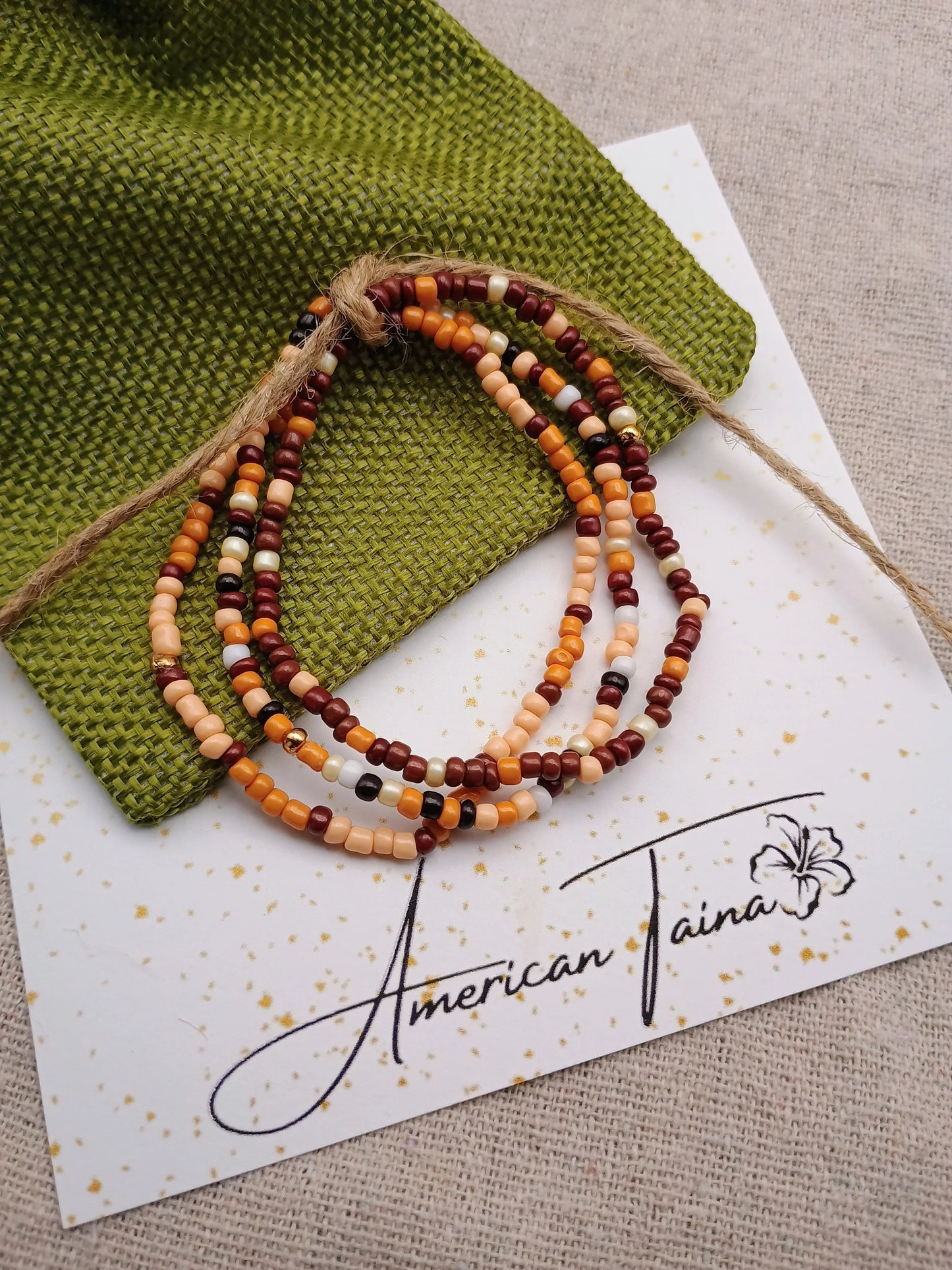 Beaded bracelet with earth tones on a green fabric pouch and white card reading American Taina