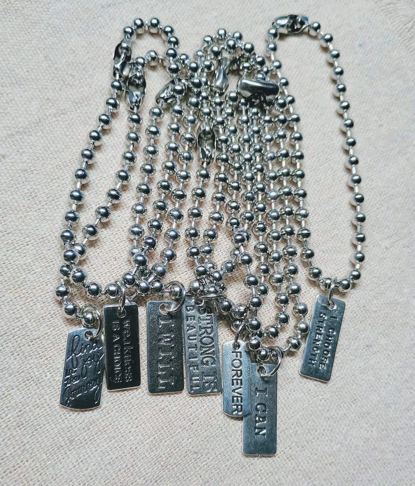 Seven silver ball chain necklaces with engraved metal tags featuring motivational phrases on beige fabric