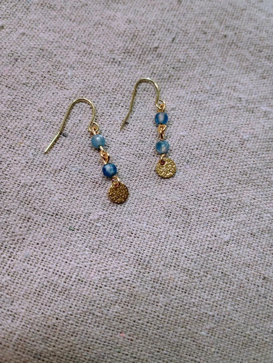 Gold hook earrings with two blue agate beads and a textured gold disc on beige fabric