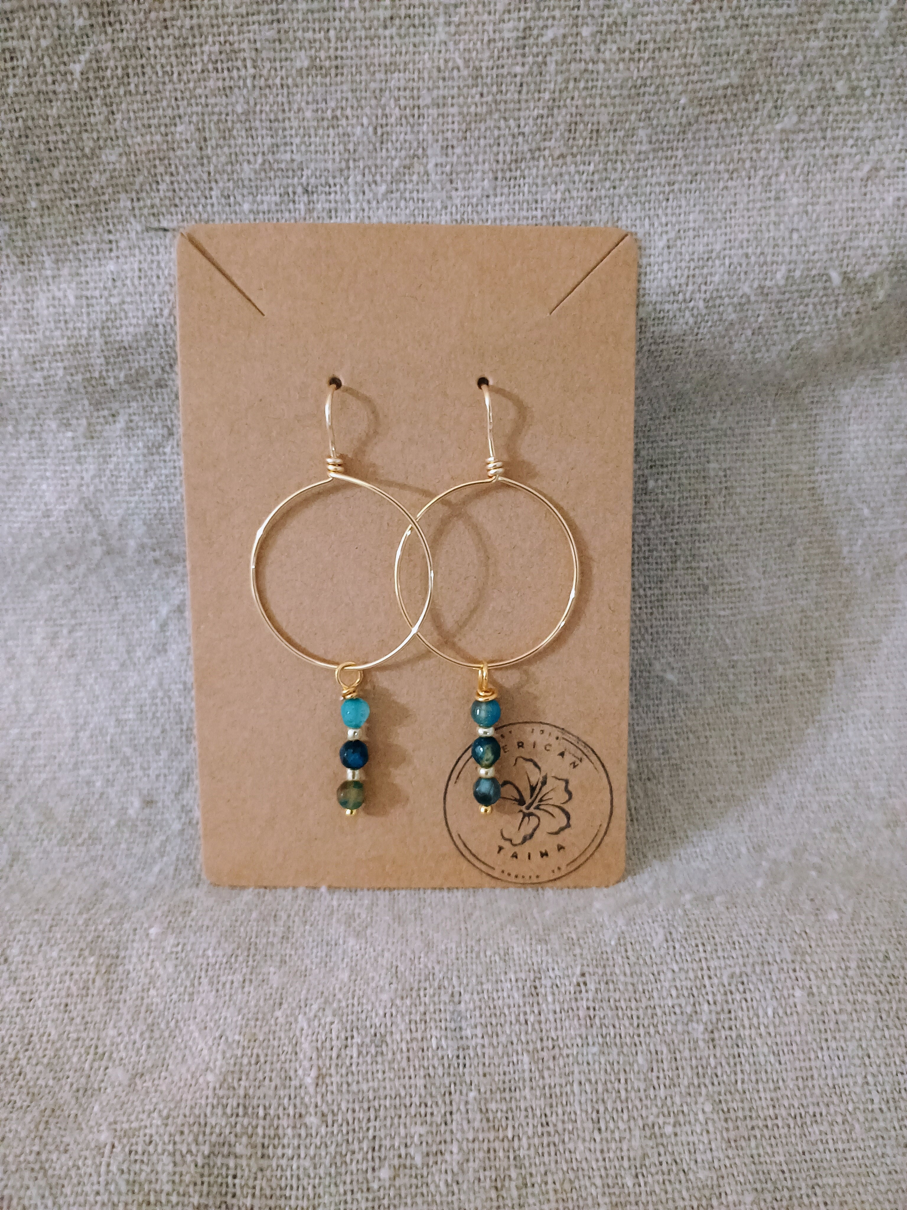 Gold hoop earrings with blue and turquoise beads on brown card against fabric background